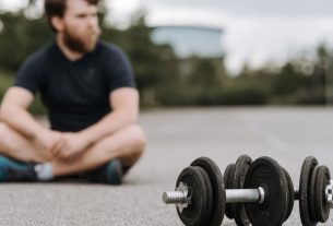 Young male with beard resting after workout on asphalt road with heavy dumbbells on gloomy day in selective focus
